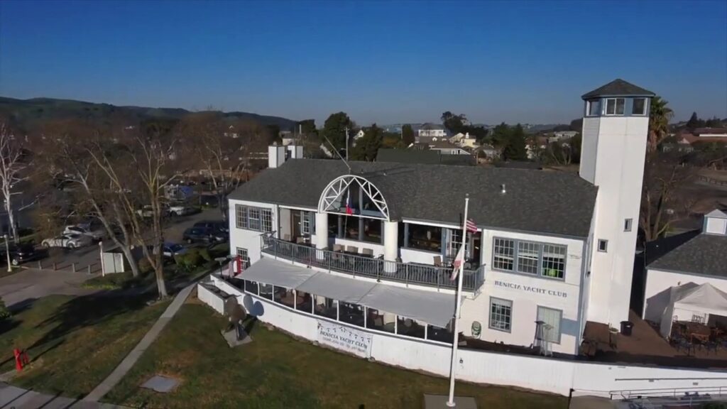 Aerial view of a two-story building with a white exterior and a round window feature, identified as a yacht club, in a suburban setting with trees and clear skies.