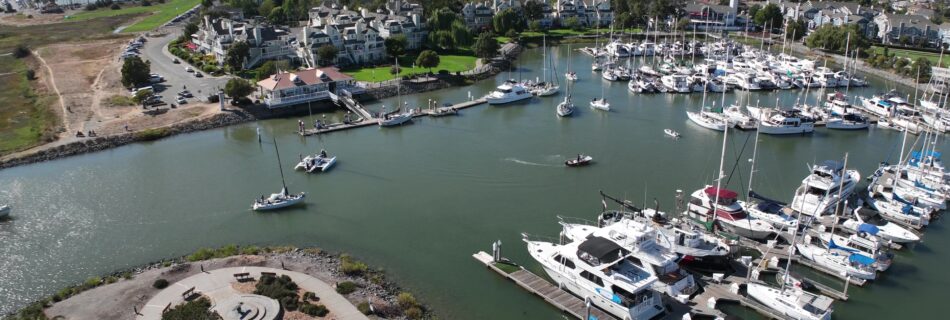 Aerial view of a marina with moored boats, adjacent to a landscaped park and residential area, with a bridge in the distance.