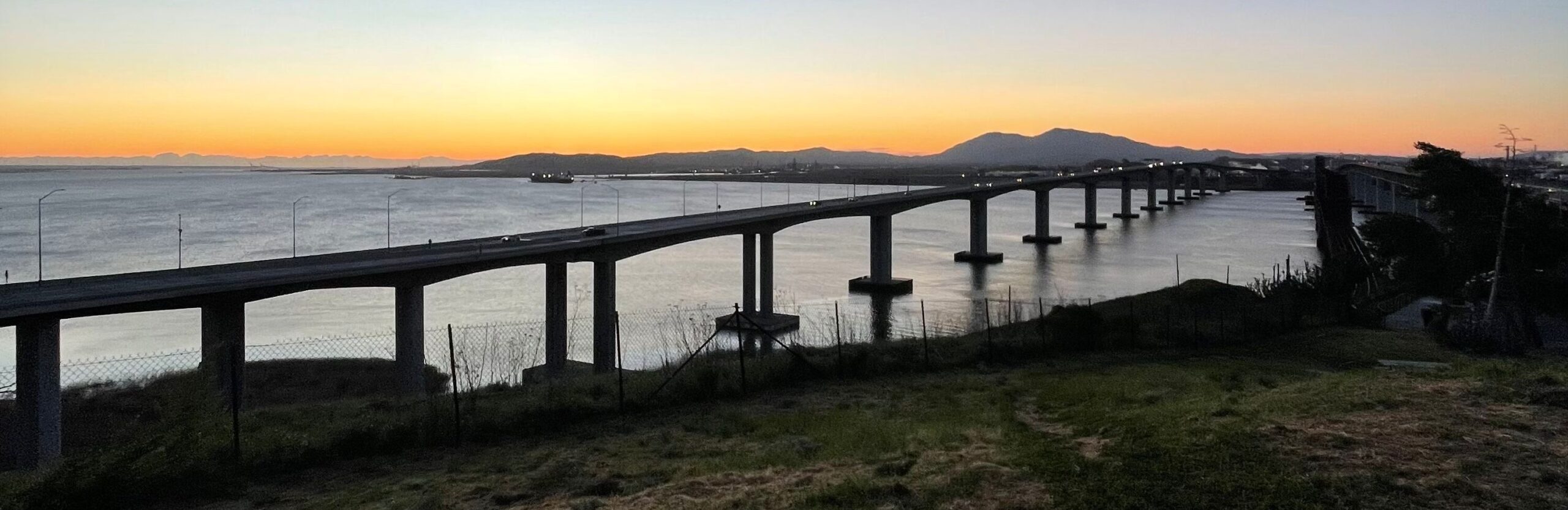 Sunset view over a coastal bridge with mountains in the distance.