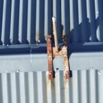 Corroded metal cross attached to a corrugated metal wall with shadows of palm leaves.