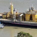 Industrial equipment and storage tanks on a river dock with boats nearby.