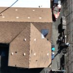 Aerial view of a two-part building with a shingled roof, a car parked alongside, and adjacent pallets beside a grassy area.