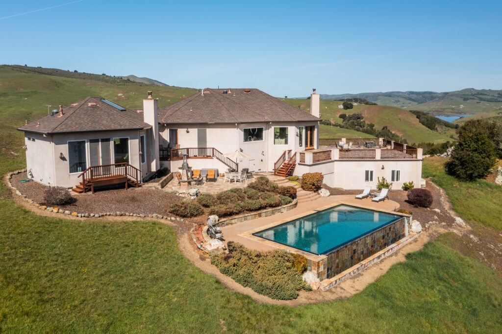 Aerial view of a large suburban home with a swimming pool, set against a rolling hillside landscape.